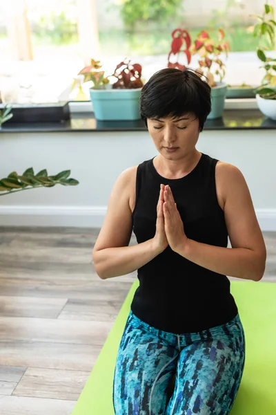 Brunette middle aged woman doing yoga meditation on exercise mat at home, mental health. Peaceful woman meditate, mental balance, time for herself, stress relief, mediation, mind fullness concept.