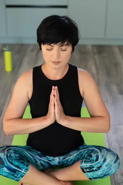 Brunette middle aged woman doing yoga meditation on exercise mat at home, mental health. Peaceful woman meditate, mental balance, time for herself, stress relief, mediation, mind fullness concept.