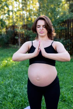 Hamile kadın dışarıda yoga yaparken nefes alıyor ve sakin, çimenler üzerinde esneme egzersizi yapıyor. Kendine Bakım, Yoga, Hamilelik, Doğum Konsepti. Anneliğin, sağlık hizmetinin, hobilerinin tadını çıkar..