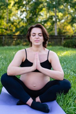 Hamile kadın dışarıda yoga yaparken nefes alıyor ve sakin, çimenler üzerinde esneme egzersizi yapıyor. Kendine Bakım, Yoga, Hamilelik, Doğum Konsepti. Anneliğin, sağlık hizmetinin, hobilerinin tadını çıkar..