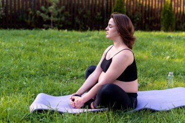 Hamile bir kadın yoga yapıyor ve bahçede çimenler üzerinde meditasyon yapıyor, temiz havayla derin bir nefes alıyor yoga nilüferleri rahat poz veriyor. Yoga Anneliği, Akıl Sağlığı ve Hamile Konsepti.