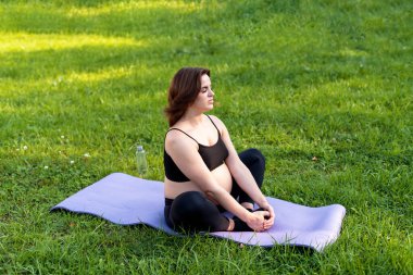 Hamile bir kadın yoga yapıyor ve bahçede çimenler üzerinde meditasyon yapıyor, temiz havayla derin bir nefes alıyor yoga nilüferleri rahat poz veriyor. Yoga Anneliği, Akıl Sağlığı ve Hamile Konsepti.