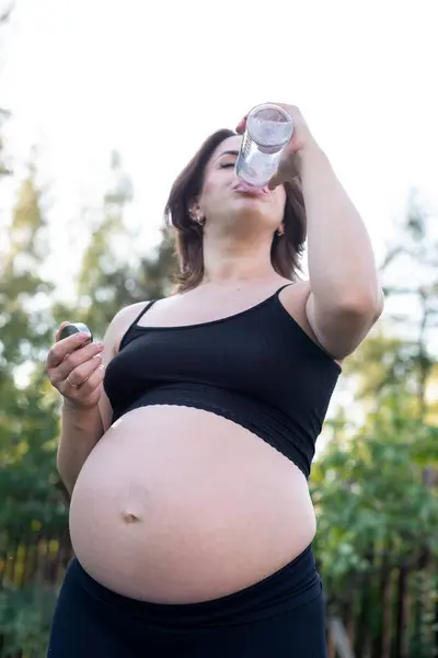 Pregnant woman in sportswear drinking a bottle of water after doing yoga outdoors, taking break in yoga practice, fitness and meditating. Motherhood, mental health and pregnancy concept.