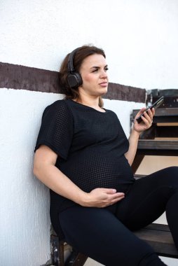 Akıllı telefonlu hamile bir kadın kulaklıkla beyaz arka planda müzik dinliyor ve boş zamanlarını dinlenerek geçiriyor. Teknoloji, annelik, akıl sağlığı, stres atma, arabuluculuk ve hamilelik.