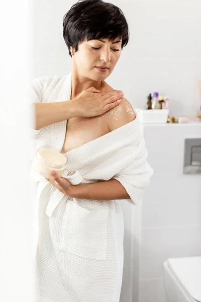 Beauty routine and body care. Cream, skincare and ethnic middle aged woman applied cosmetic moisturizing cream lotion on her body in bathroom. Beauty treatment at home