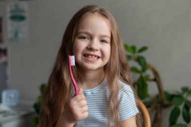 Beyaz, genç bir kız. Dişi kopmuş, gülümsüyor ve elinde pembe diş fırçası var..