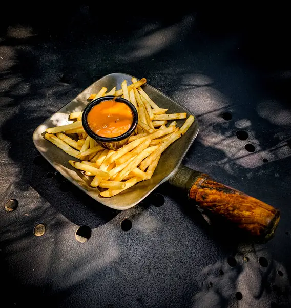 Lezzetli portakal soslu çıtır patates kızartmasının yaratıcı bir sunumu. Koyu dokulu bir yüzeyde eşsiz bir kürek stili tabakta servis ediliyor. Sanatsal gölgeler yaratıyor..