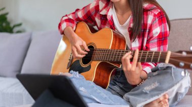 Concept of relaxation with music, Young woman plays acoustic guitar while learning music on tablet.