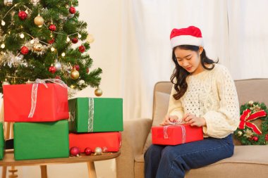 Noel kutlaması konsepti, genç Asyalı kadın koltukta oturuyor ve noel hediyesini açıyor..