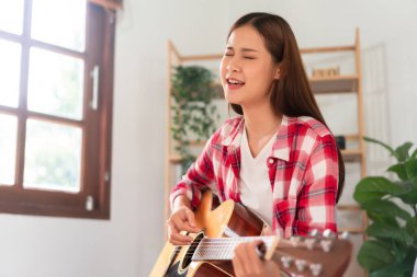 Hobi olarak, genç Asyalı kadın oturma odasında dinlenmek için akustik gitar çalıp şarkı söylüyor..