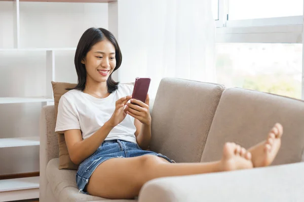 stock image Cozy lifestyle concept, Young woman lying on couch and use smartphone to surf social media at home.