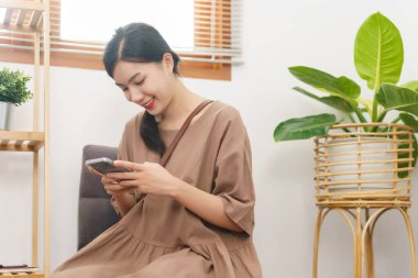 Rahatlama yaşam tarzı konsepti, genç Asyalı kadın oturma odasında sosyal medyada sörf yapmak için akıllı telefon kullanıyor..