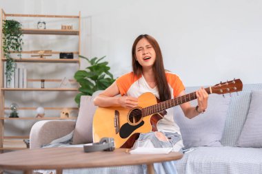 Hobi konsepti, genç Asyalı kadın kanepede oturup akustik gitarla şarkı söylemek ve müzik çalmak..