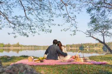 Genç aşık bir çift piknik sepetiyle battaniyeye oturmuş göl manzarasını seyrediyor..