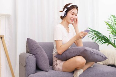 Yaşam tarzı oturma odası konsepti, kulaklıklı genç Asyalı kadın akıllı telefondan müzik dinliyor..