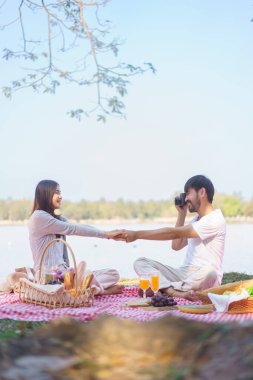 Genç aşıklar el ele tutuşup fotoğraf çekerken piknik için battaniyeye otururlar..