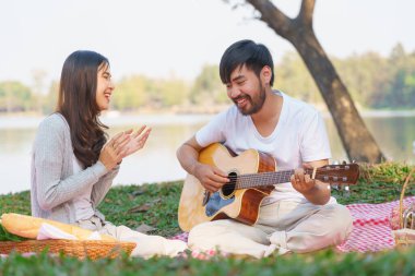 Kız arkadaşı için akustik gitar çalan genç bir adam piknik yapmak için battaniyeye oturmuş..