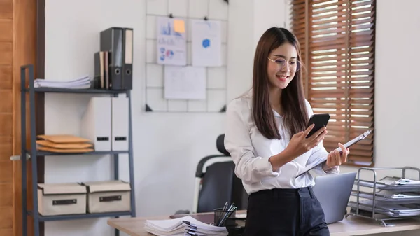 Business concept, Businesswoman holding clipboard and stand to reading business data in smartphone.