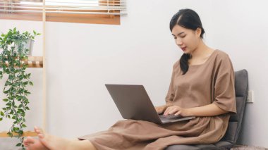 Rahatlama yaşam tarzı konsepti, oturma odasında sosyal medyada sörf yapmak için dizüstü bilgisayar kullanan genç Asyalı kadın..