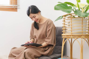 Rahatlama yaşam tarzı konsepti, oturma odasında sosyal medyada sörf yapmak için tablet kullanan genç Asyalı kadın..