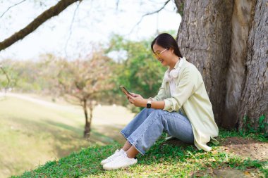Kadınlar çimenlerde oturup akıllı telefondan sosyal medyada sörf yaparken parkta ağaçların altında dinleniyorlar..