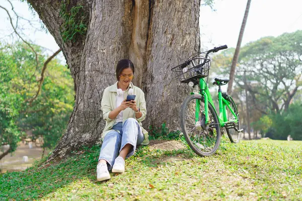 stock image Women sitting under tree and surfing social media on smartphone to resting after cycling in park.