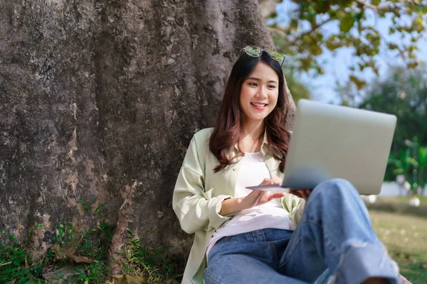 Kadınlar, parkta dinlenirken dizüstü bilgisayarlarında çalışmak ve sosyal medyada sörf yapmak için ağaca yaslanıyorlar..