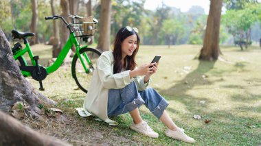 Ağaçların altında oturan kadınlar parkta bisiklet sürdükten sonra sosyal medyada çalışmak ve sörf yapmak için akıllı telefon kullanıyorlar..