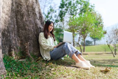 Kadınlar, parkta dinlenirken dizüstü bilgisayarlarında çalışmak ve sosyal medyada sörf yapmak için ağaca yaslanıyorlar..