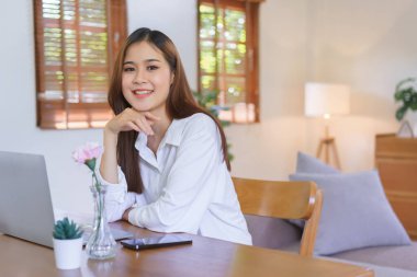 Rahatlatıcı zaman kavramı, modern ev ofisinde çalışırken gülümseyen ve kameraya bakan kadın..