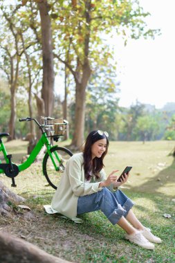 Ağaçların altında oturan kadınlar parkta bisiklet sürdükten sonra sosyal medyada çalışmak ve sörf yapmak için akıllı telefon kullanıyorlar..