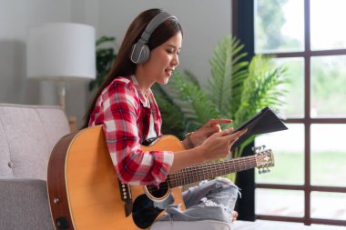 Hobi konsepti, genç Asyalı kadın kulaklık takıyor ve tablette müzik öğrenmek için kanepeye oturuyor..