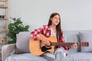 Gitar ve şarkıcı konsepti, genç Asyalı kadın kanepede şarkı söylemek için akustik gitar çalıyor..