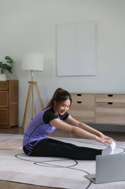 Genç Asyalı kadın antrenman için dizüstü bilgisayarda spor videosu izliyor ve bacak esnetme egzersizi yapıyor..
