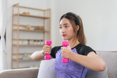 Kanepede oturan genç Asyalı kadın güçlü kolları çalıştırmak için halterleri kaldırarak egzersiz yapıyor..