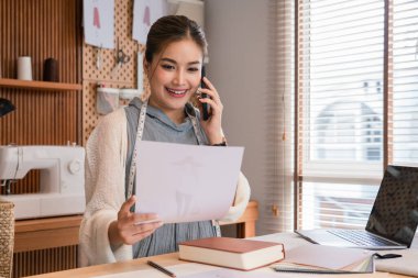 Akıllı telefondan müşteriyle konuşan Asyalı terzi kadınlar ve belgelerdeki kıyafet detaylarını kontrol ediyorlar.