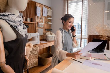 Akıllı telefondan müşteriyle konuşan Asyalı terzi kadınlar ve belgelerdeki kıyafet detaylarını kontrol ediyorlar.