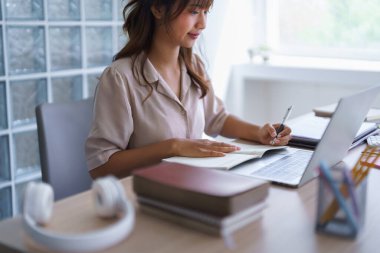 Gündelik giysiler içinde Asyalı genç bir öğrenci öğretim görevlileri eğitimini online olarak bilgisayar teknolojisiyle okurken aynı zamanda bilgi verileri yazıp üniversitede ödev yapıyor..