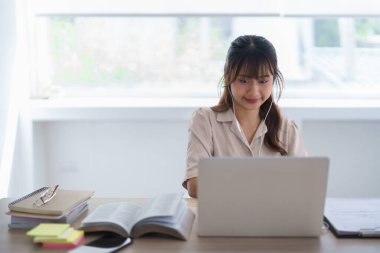 Gündelik kıyafetlerle dolaşan Asyalı genç bir öğrenci, özel ders izlemek ve ders çalışmak için üniversitede bilgi verileri yazarken ve ödevlerini yaparken online ders çalışmak için kulaklık takıyor..