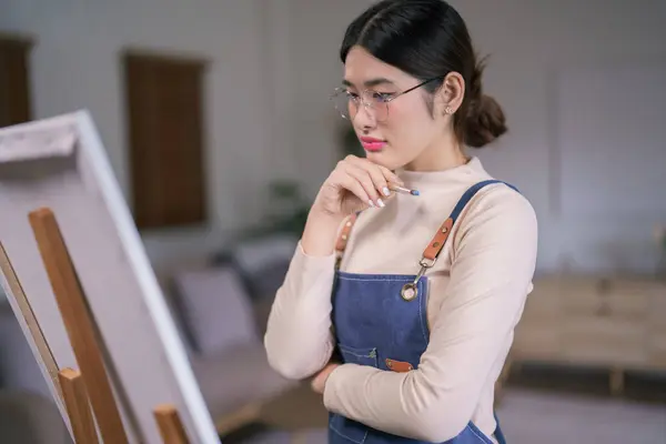 若いアジアの女性アーティストは絵画のアートワークを作成することを考えるためにペンキブラシとキャンバスを見ています — ストック写真