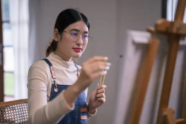 Young asian female artist hold paintbrush and use brush to drawing and painting artwork on canvas.