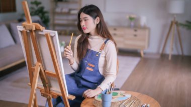 Genç Asyalı kadın sanatçı, resim yapmak için tuval üzerindeki orantıyı ölçmek üzere kalem tutuyor..