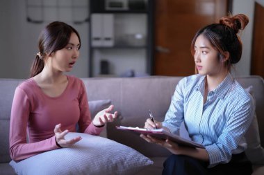 Stresli Asyalı hasta kadınlar doktorlara psikolojik sağlık sorunlarını açıklarken kadın psikoloğlar not panosuna veri notları ve zihinsel sağlık terapisi hakkında danışmanlık alıyorlar..