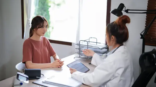 Asian psychologist women reading health examination results and writing notes on tablet while giving counseling to explaining about medicine with mental health therapy to female patient in clinic.