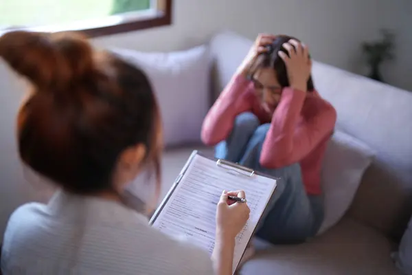 Asian patient women stressed and headache with anxiety while female psychologist examining about psychological health problem and writing notes on paper to counseling about mental health therapy.