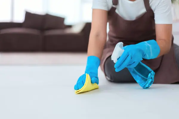 Maid wearing gloves and using cleaner spray and microfiber cloth to wipe a dust on the floor.