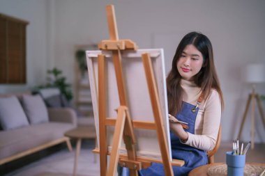 Genç Asyalı kadın sanatçı tuval üzerinde resim yapmak için renk paletiyle boya fırçası kullanıyor..