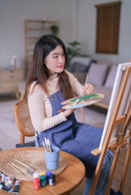 Genç Asyalı kadın sanatçı tuval üzerinde resim yapmak için palet üzerindeki renkleri karıştırmak için boya fırçası kullanıyor.