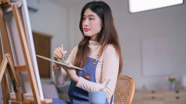 stock image Young asian female artist using paintbrush to mixing color on palette to create painting on canvas.