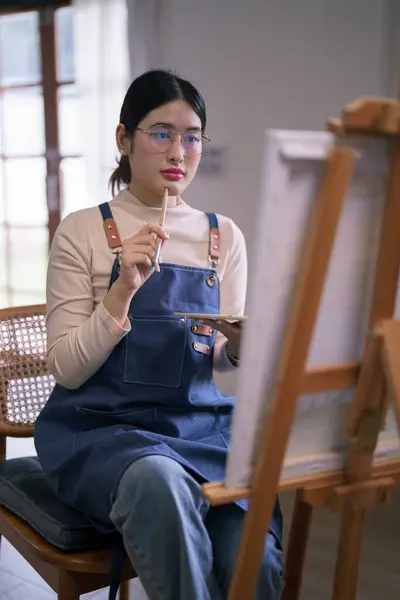 Young asian female artist hold paintbrush while looking on canvas to thinking idea painting artwork.
